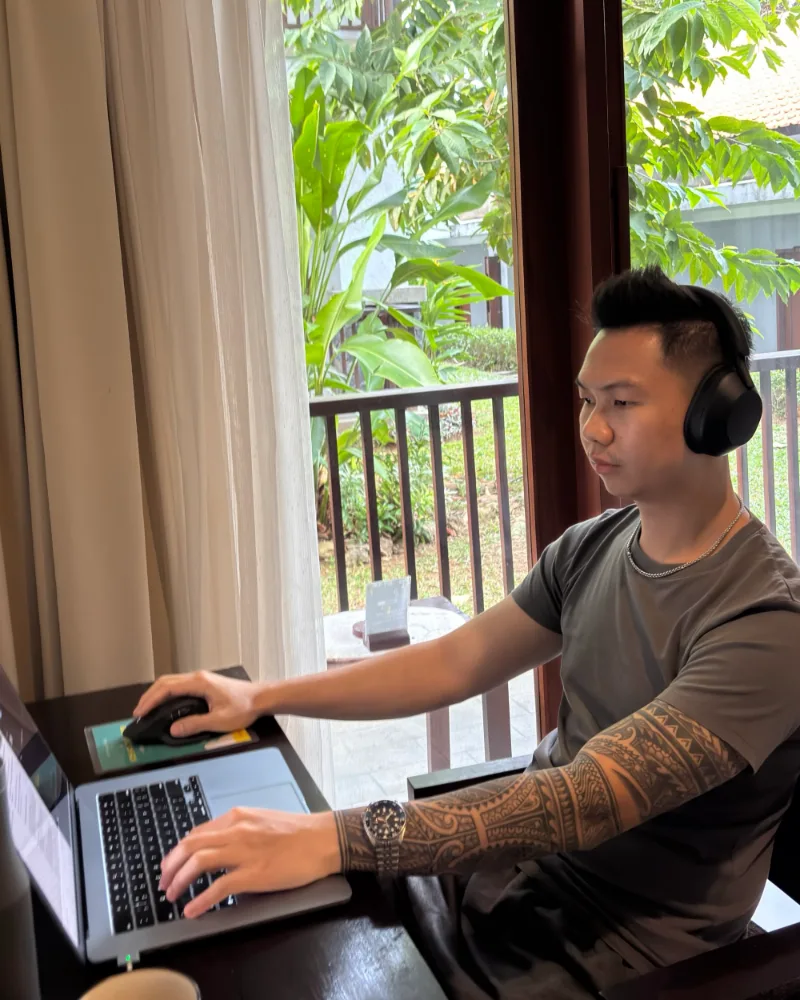 Gue lagi kerja di depan laptop pakai headset, di samping jendela dengan latar taman hijau.