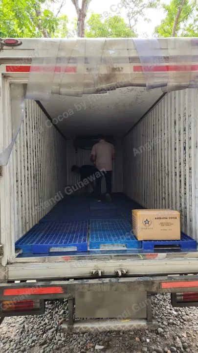 Bak truk boks terbuka dengan palet biru di lantainya dan satu kardus Angkassa Cocoa, dua orang menata barang di dalam.